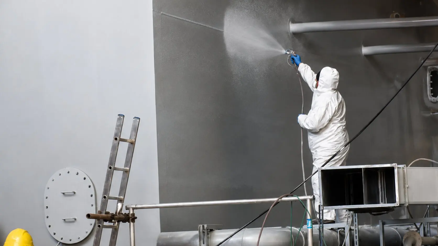Operator in protective suit and respirator spray-applying the coating to a steel structure from a scaffold in the workshop