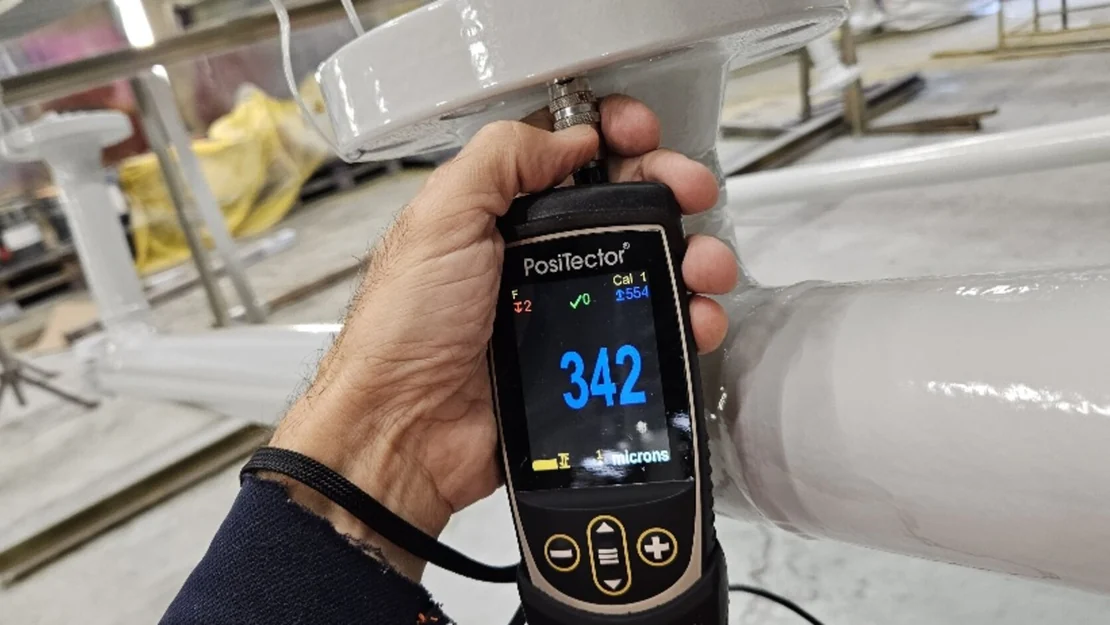 A hand holding a digital thickness gauge against a coated pipeline: the display reads 342 microns dry film thickness